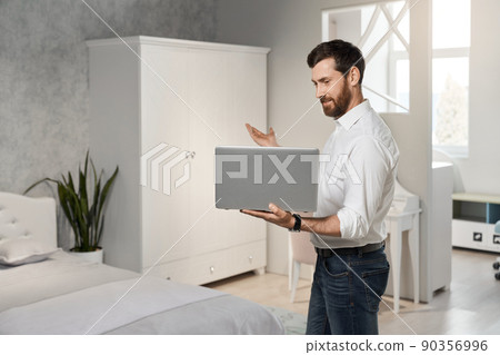 Side view of handsome seller, designer, architect standing, holding laptop. Brunette male with beard looking at screen, showing, representing bedroom. Concept of designing. Side view of handsome seller, designer, architect standing, holding laptop. Brunette male with beard looking at screen, showing, representing bedroom. Concept of designing. 90356996