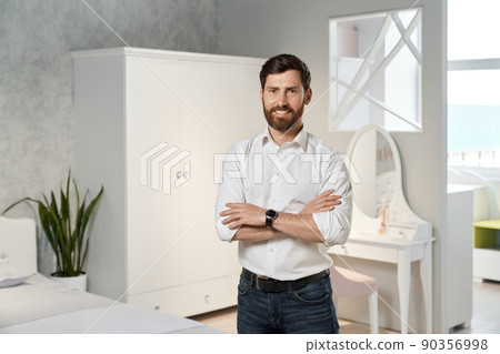 Front view of handsome seller, designer, architect standing with crossed hands in bedroom. Brunette male with beard designing, looking at camera, smiling. Concept of furniture. Front view of handsome seller, designer, architect standing with crossed hands in bedroom. Brunette male with beard designing, looking at camera, smiling. Concept of furniture. 90356998