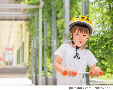 Little boy rides kick scooter in skate park. Portrait of kid in helmet on green leaves background. Training to skate at summer. Little boy rides kick scooter in skate park. Portrait of kid in helmet on green leaves background. Training to skate at summer. 90357652