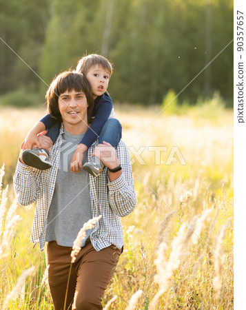 Father carries his son on shoulders. Family time outdoors. Man with child on autumn field at sunset. Dad and son explores nature together. Father carries his son on shoulders. Family time outdoors. Man with child on autumn field at sunset. Dad and son explores nature together. 90357657