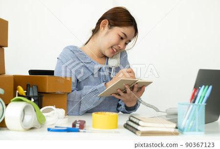 Young Asian woman business owner with many parcel boxes on the table happy online sales job, use the laptop and telephone, get an order from customers, take notes 90367123