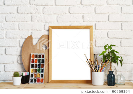 Photo frame as easel with artist's tools on wooden table against white brick wall Photo frame as easel with artist's tools on wooden table against white brick wall 90369394