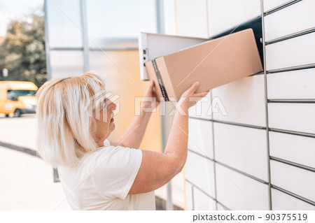 50s caucasian woman getting parcel from cell of automatic post terminal outdoors.  90375519