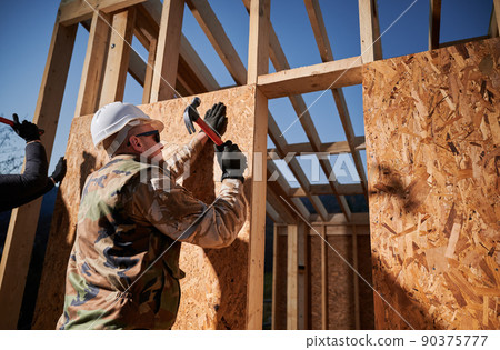 Carpenter hammering nail into OSB panel on the wall of future cottage. Man worker building wooden frame house in the Scandinavian style barnhouse. Carpentry and construction concept. Carpenter hammering nail into OSB panel on the wall of future cottage. Man worker building wooden frame house in the Scandinavian style barnhouse. Carpentry and construction concept. 90375777