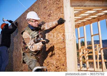 Carpenters hammering nail into OSB panel on the wall of future cottage. Men workers building wooden frame house. Carpentry and construction concept. Carpenters hammering nail into OSB panel on the wall of future cottage. Men workers building wooden frame house. Carpentry and construction concept. 90375782