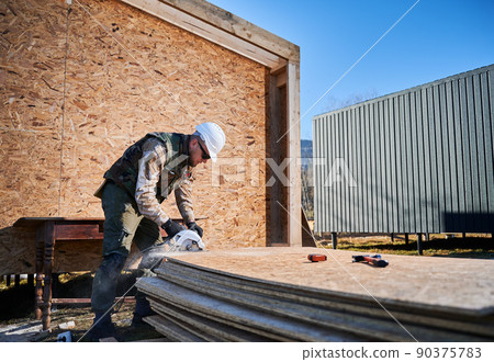 Carpenter using circular saw for cutting wooden OSB board. Man worker building wooden frame house on pile foundation. Carpentry concept. Carpenter using circular saw for cutting wooden OSB board. Man worker building wooden frame house on pile foundation. Carpentry concept. 90375783
