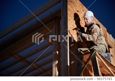 Carpenter hammering nail into OSB panel on the wall of future cottage at night. Man worker building wooden frame house. Carpentry and construction concept. 90375785