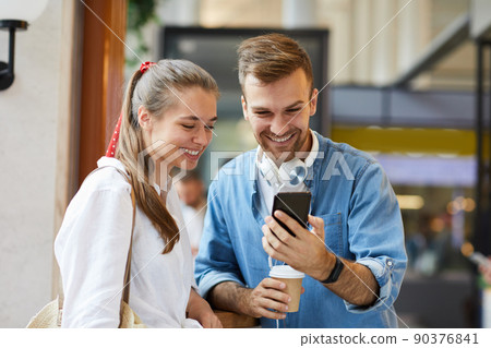 Cheerful young friends in casual clothing standing in cafe and watching online video on smartphone 90376841