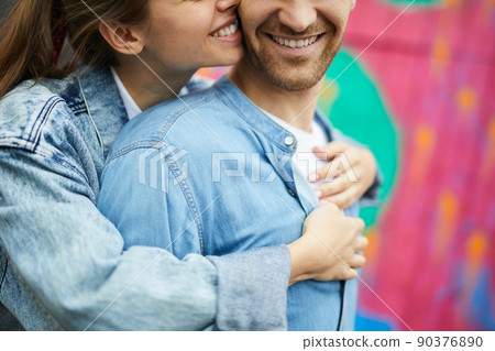 Close-up of happy affectionate woman in denim jacket cuddling boyfriend from back and making sounds in his ear 90376890