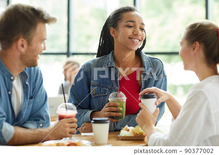 Smiling attractive young Black woman in denim jacket standing at table and enjoying time with friends 90377002