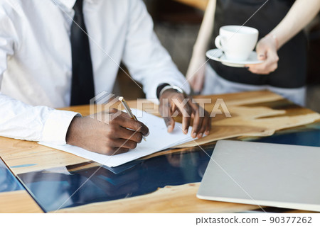 Close-up of busy black businessman sitting at wooden table in restaurant and examining documents while waitress brining coffee to him 90377262