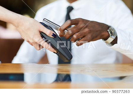 Close-up of unrecognizable black businessman with elegant wristwatch paying for bill with smartphone in cafe, waitress holding terminal with nfc 90377263