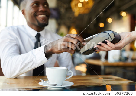 Close-up of positive young African-American businessman sitting at table and using smartphone while making contactless payment in cafe Close-up of positive young African-American businessman sitting at table and using smartphone while making contactless payment in cafe 90377266