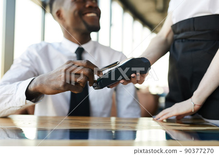 Close-up of smiling black businessman sitting at table and using nfc technology for mobile payment in restaurant Close-up of smiling black businessman sitting at table and using nfc technology for mobile payment in restaurant 90377270