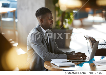 Serious busy young African-American businessman with beard sitting at table and typing on laptop while browsing internet 90377363