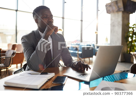 Serious thoughtful young black businessman in gray suit sitting at wooden table and browsing internet on laptop while taking information for report Serious thoughtful young black businessman in gray suit sitting at wooden table and browsing internet on laptop while taking information for report 90377366