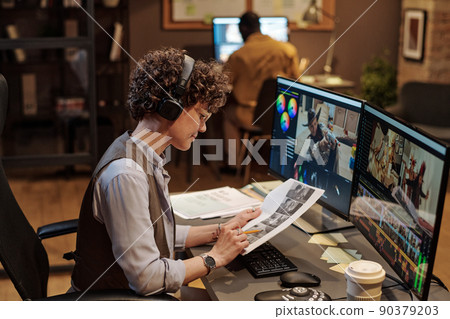 Female editor in headphones examining script at her workplace while editing photos on computer 90379203