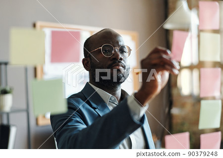 Serious African businessman in suit and eyeglasses making notes on sticky notes hanging on glass wall at office 90379248