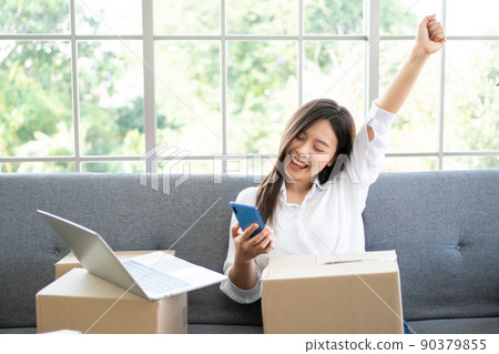 Happy young Asian woman entrepreneur, Smile for sales success after checking order from online shopping store in a smartphone at home office, Concept of merchant business online and eCommerce 90379855