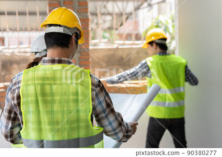 Asian engineer or Young Architect put on a helmet for safety and look at Blueprint for Inspect Building factory Construction Site and use walkie talkie for a talk with a contractor. 90380277