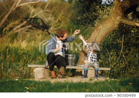 Young dad and his child boy spend time outdoors together. Curly toddler boy wearing a plaid shirt and a hat. Boy sitting near the tree and listening his father. Young dad and his child boy spend time outdoors together. Curly toddler boy wearing a plaid shirt and a hat. Boy sitting near the tree and listening his father. 90380499
