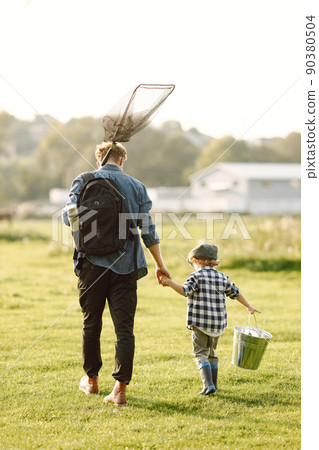 Dad and his child boy spend time outdoors together. Curly toddler boy wearing a plaid shirt and a hat. Boy holding a hand of his father. 90380504