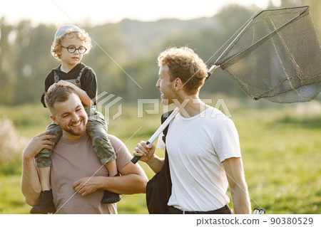 Dad, his friend and his child boy spend time outdoors together. Curly toddler boy wearing a khaki overall. Boy sitting on a shoulders of his father. 90380529