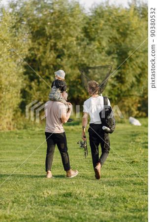 Dad, his friend and his child boy spend time outdoors together. Curly toddler boy wearing a khaki overall. Boy sitting on a shoulders of his father. 90380532