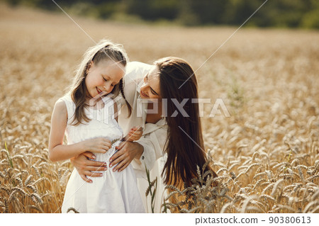 ute photo of a young mother and her daughter in white dresses at the wheat field on a sunny day. Brunette mother and her little daughter posing for a photo. Girl looking at wheat spike. 90380613