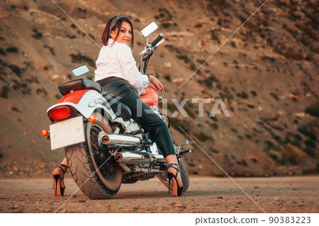 A beautiful adult woman with a high heels and leather pants, sexy posing sitting on motorcycle. Rear view. Rock on the background. The concept of Motorcyclist Day A beautiful adult woman with a high heels and leather pants, sexy posing sitting on motorcycle. Rear view. Rock on the background. The concept of Motorcyclist Day 90383223