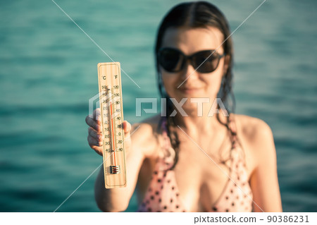 Beautiful young woman in sunglasses with a thermometer in her hand on the background of the sea. Summer vacation and heat concept, selective focus 90386231