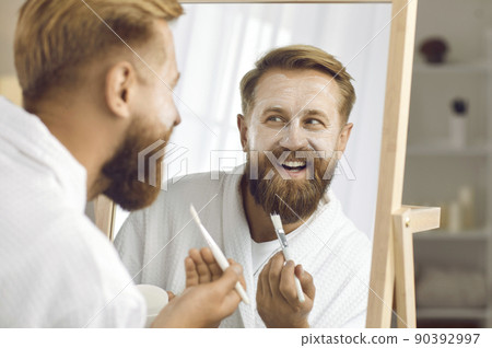 Close up of funny man reflected in mirror who applies cosmetic mask for facial care. Close up of funny man reflected in mirror who applies cosmetic mask for facial care. 90392997