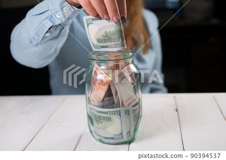 Close-up of a young woman putting hundred-dollar bills in a glass jar. Close-up of a young woman putting hundred-dollar bills in a glass jar. 90394537