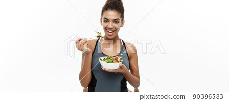 Healthy and Fitness concept - Beautiful American African lady in fitness clothes on diet eating fresh salad. Isolated on white background. Healthy and Fitness concept - Beautiful American African lady in fitness clothes on diet eating fresh salad. Isolated on white background. 90396583
