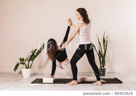 Two women in sportswear practicing yoga doing a low lunge on one leg with a twist or Anjanasana or Anjaneyasana (Crescent Pose or Young Moon Pose). The teacher helps the student 90403829