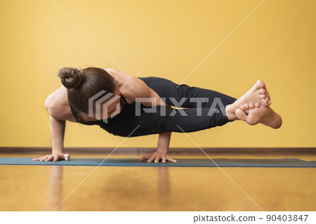 A woman in a one-piece black tracksuit performs the Ashtavakrasana exercise, a handstand against the background of the wall 90403847