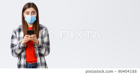 Different emotions, covid-19, social distancing and technology concept. Surprised young woman got news via text message, raising head from mobile phone and stare camera impressed, wear medical mask 90404188