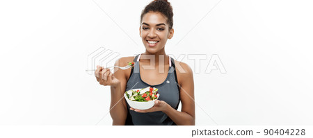 Healthy and Fitness concept - Beautiful American African lady in fitness clothes on diet eating fresh salad. Isolated on white background. Healthy and Fitness concept - Beautiful American African lady in fitness clothes on diet eating fresh salad. Isolated on white background. 90404228