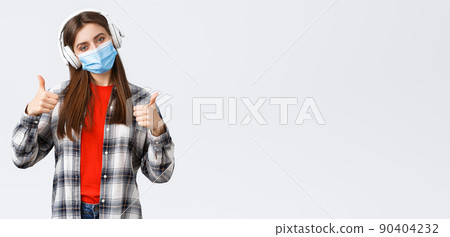 Social distancing, leisure and lifestyle on covid-19 outbreak, coronavirus concept. Pleased good-looking woman in medical mask, listening music headphones, show thumb-up good headsets Social distancing, leisure and lifestyle on covid-19 outbreak, coronavirus concept. Pleased good-looking woman in medical mask, listening music headphones, show thumb-up good headsets 90404232
