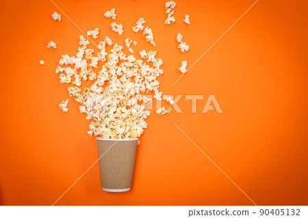 Popcorn spills out of the glass on an orange background, viewed from above. A splash of popcorn Popcorn spills out of the glass on an orange background, viewed from above. A splash of popcorn 90405132