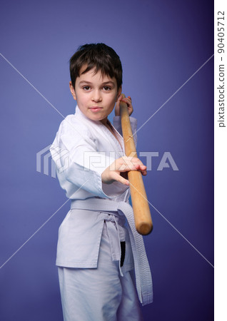 Confident strong concentrated boy - Aikido wrestler - in a kimono stands with a wooden jo weapon in his hands. Oriental martial arts practice concept Confident strong concentrated boy - Aikido wrestler - in a kimono stands with a wooden jo weapon in his hands. Oriental martial arts practice concept 90405712