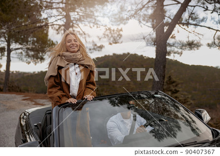 Beautiful Caucasian young woman standing on seat in convertible car with husband and smiling 90407367