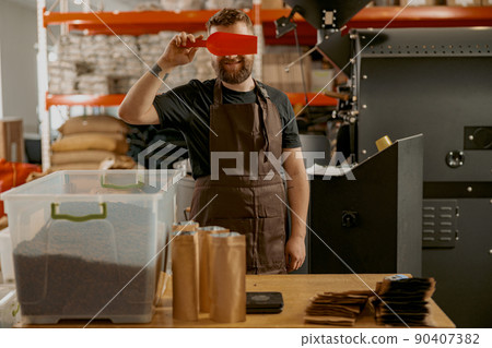 Playful barista closes eyes with a small ladle while standing at a small coffee factory Playful barista closes eyes with a small ladle while standing at a small coffee factory 90407382