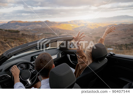 Rear of happy couple man and woman traveling in convertible car by mountainside enjoying view 90407544