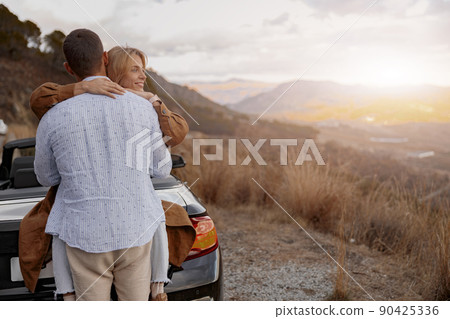 Rear of a man hugging his girlfriend outdoors at sunset, mountains view, love relationship 90425336