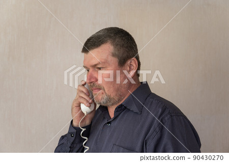A portrait of a grown man talking on a corded phone. A man in a blue striped shirt is holding a white telephone receiver next to his head. Short brown hair with gray. 90430270