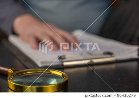 DEFOCUSED SHOT of a hand resting on a clipboard. A gold-rimmed magnifying glass is in the foreground. Adult male laying his hand on documents with handwritten text. Blur, noise. Close-up. Selective DEFOCUSED SHOT of a hand resting on a clipboard. A gold-rimmed magnifying glass is in the foreground. Adult male laying his hand on documents with handwritten text. Blur, noise. Close-up. Selective 90430279