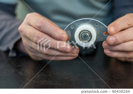 A grown man holds in his lower roller for a sliding door. Metal mounting plate with white plastic roller. Furniture hardware. Close-up. Selective focus. A grown man holds in his lower roller for a sliding door. Metal mounting plate with white plastic roller. Furniture hardware. Close-up. Selective focus. 90430284