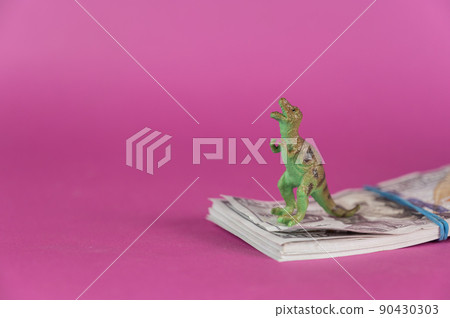 A dinosaur and a wad of cash against a purple background. A stack of hundred-dollar bills. Green miniature of a predatory animal standing on its hind legs. Selective focus. A dinosaur and a wad of cash against a purple background. A stack of hundred-dollar bills. Green miniature of a predatory animal standing on its hind legs. Selective focus. 90430303