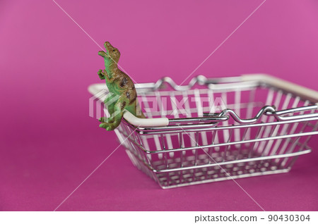 Dinosaur and shopping cart against a purple background. The dinosaur sits on a white handle. Green miniature of an animal of prey. Selective focus. Dinosaur and shopping cart against a purple background. The dinosaur sits on a white handle. Green miniature of an animal of prey. Selective focus. 90430304